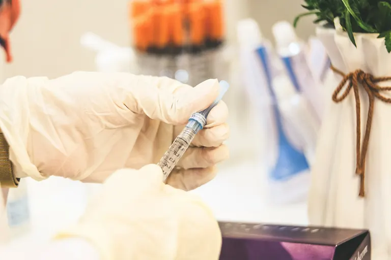 Gloved hands holding a graduated syringe in a clinic treatment room, with skincare products on shelves behind