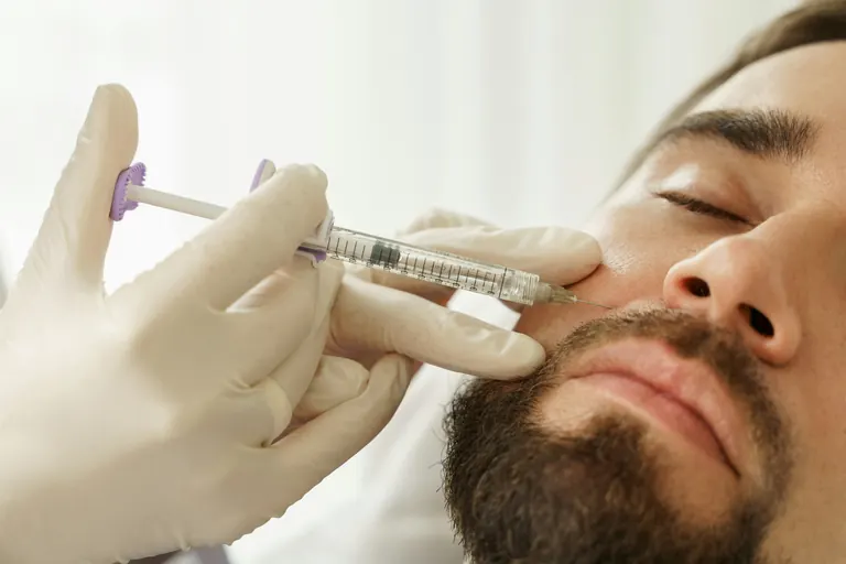 A gloved clinician injecting the cheek of a reclining bearded man with a fine needle