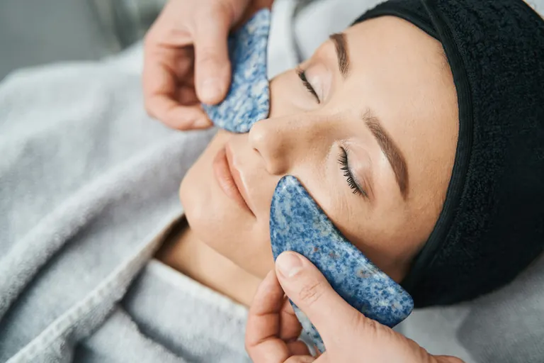 Cool blue stone tools held against a reclining woman’s cheeks by gloved hands, her eyes closed and a towel around her hair
