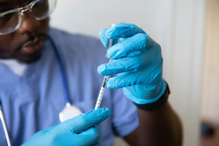 A clinician in scrubs and gloves drawing solution from a vial into a graduated syringe