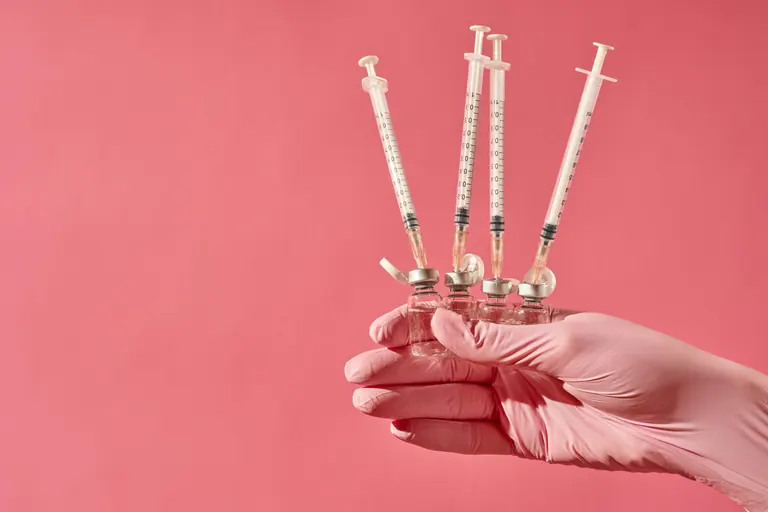 A gloved hand holding four prepared syringes fanned out against a pink background