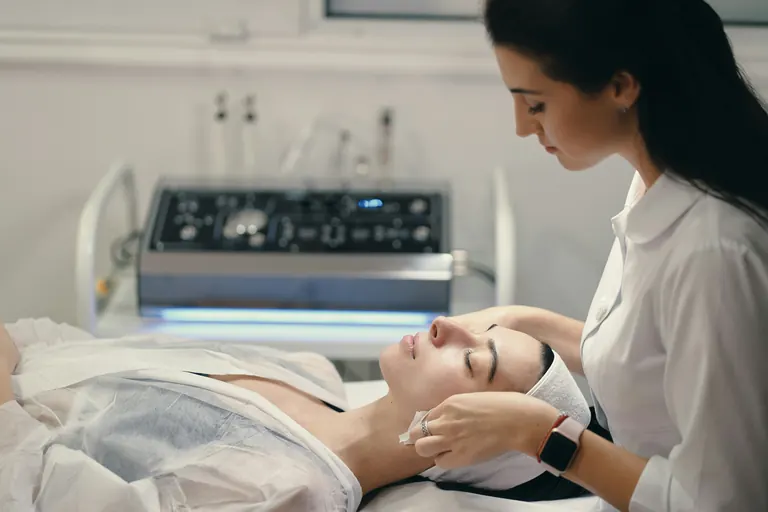 A patient receiving a device-based facial treatment in a clinic room with equipment behind