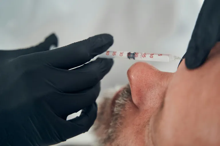A graduated syringe held horizontally beside a man’s nose by black-gloved hands, unit markings visible along the barrel