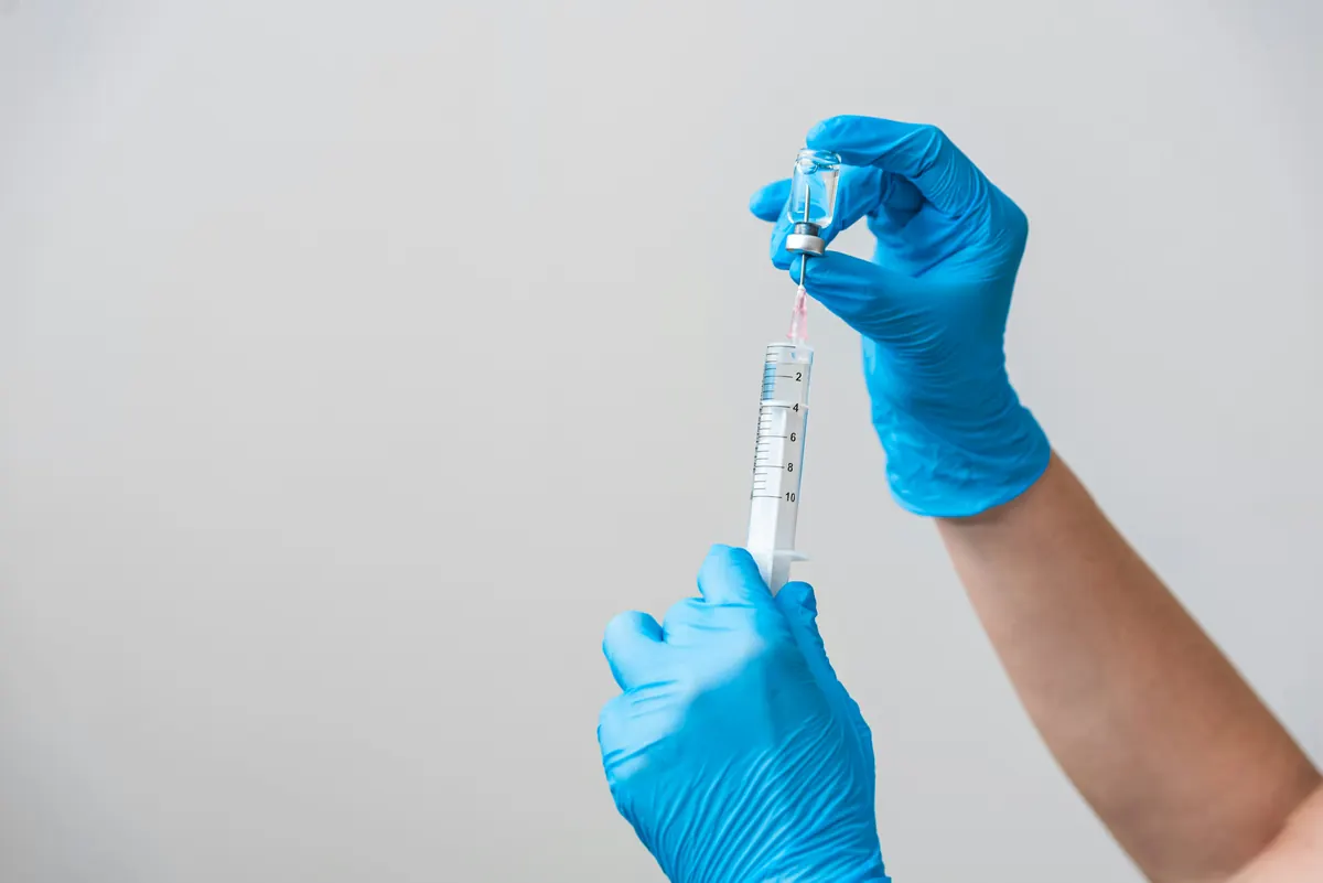 A clinician in white gloves holding a small glass vial and drawing its contents into a syringe