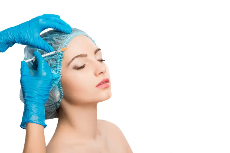 A gloved clinician placing an injection near the outer brow of a patient wearing a surgical cap, against a plain white background