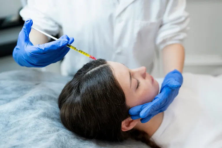 A gloved clinician holding a graduated syringe above a patient lying on a treatment couch, steadying her head with the other hand