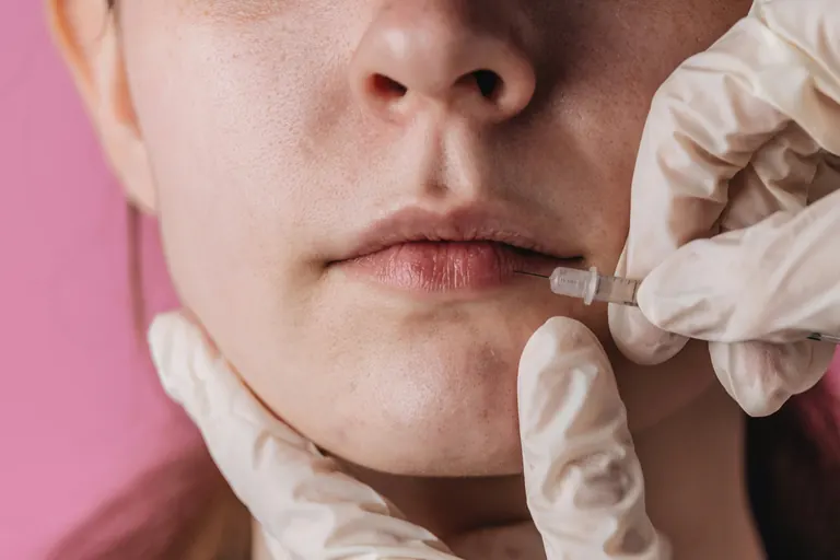 A small syringe positioned beside a woman’s mouth, her lower face filling the frame against a pink background