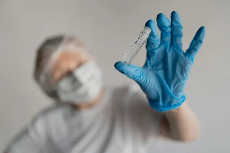 A masked person in a blue glove holding up a small unlabelled glass vial