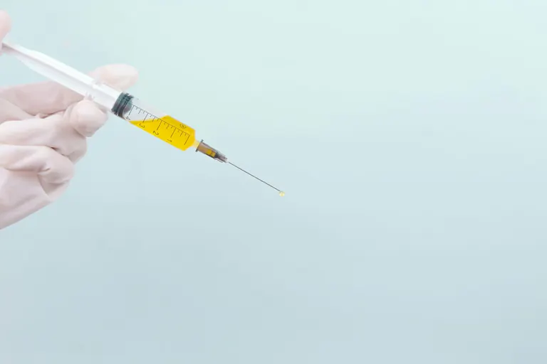 A gloved hand holding a graduated syringe of pale yellow solution against a plain background, with a droplet at the needle tip