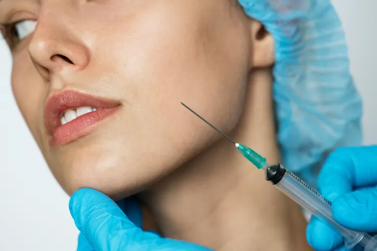 A fine needle approaching the jawline below the corner of a woman’s mouth, held by a gloved clinician