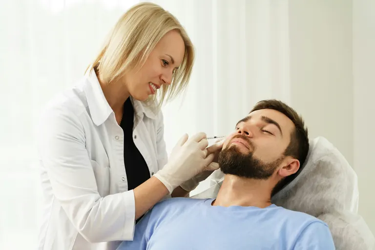 A clinician in a white coat injecting the lower face and jaw area of a reclining male patient in a treatment room