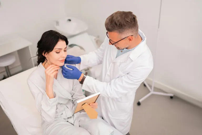 A clinician in a white coat holding a clipboard examining a seated patient in a clinical treatment room