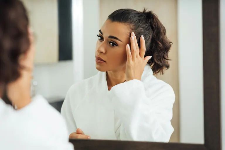A woman in a white robe studying her own face closely in a mirror, one hand raised to her temple
