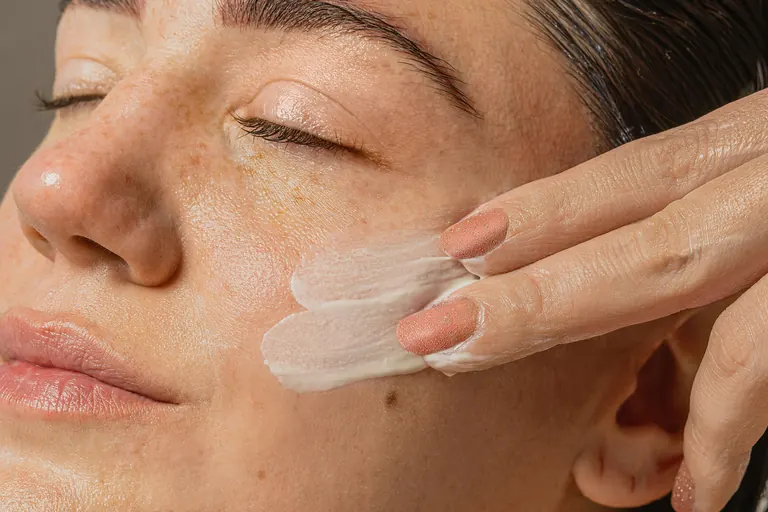 A close-up of a woman&rsquo;s cheek as a pale cream is smoothed across it with two fingertips, with the skin texture and pores clearly visible