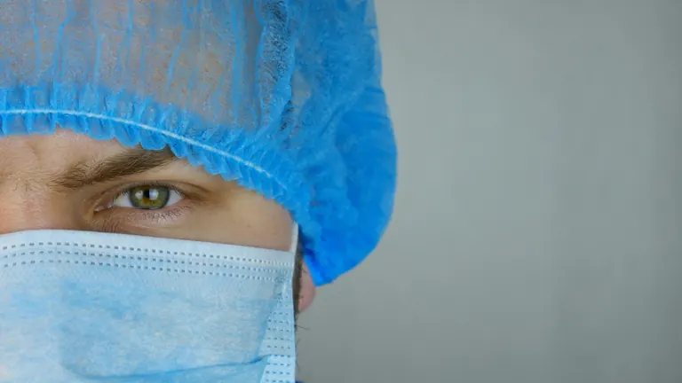 A close-up of a clinician in a surgical cap and mask, one eye visible, against a plain grey background